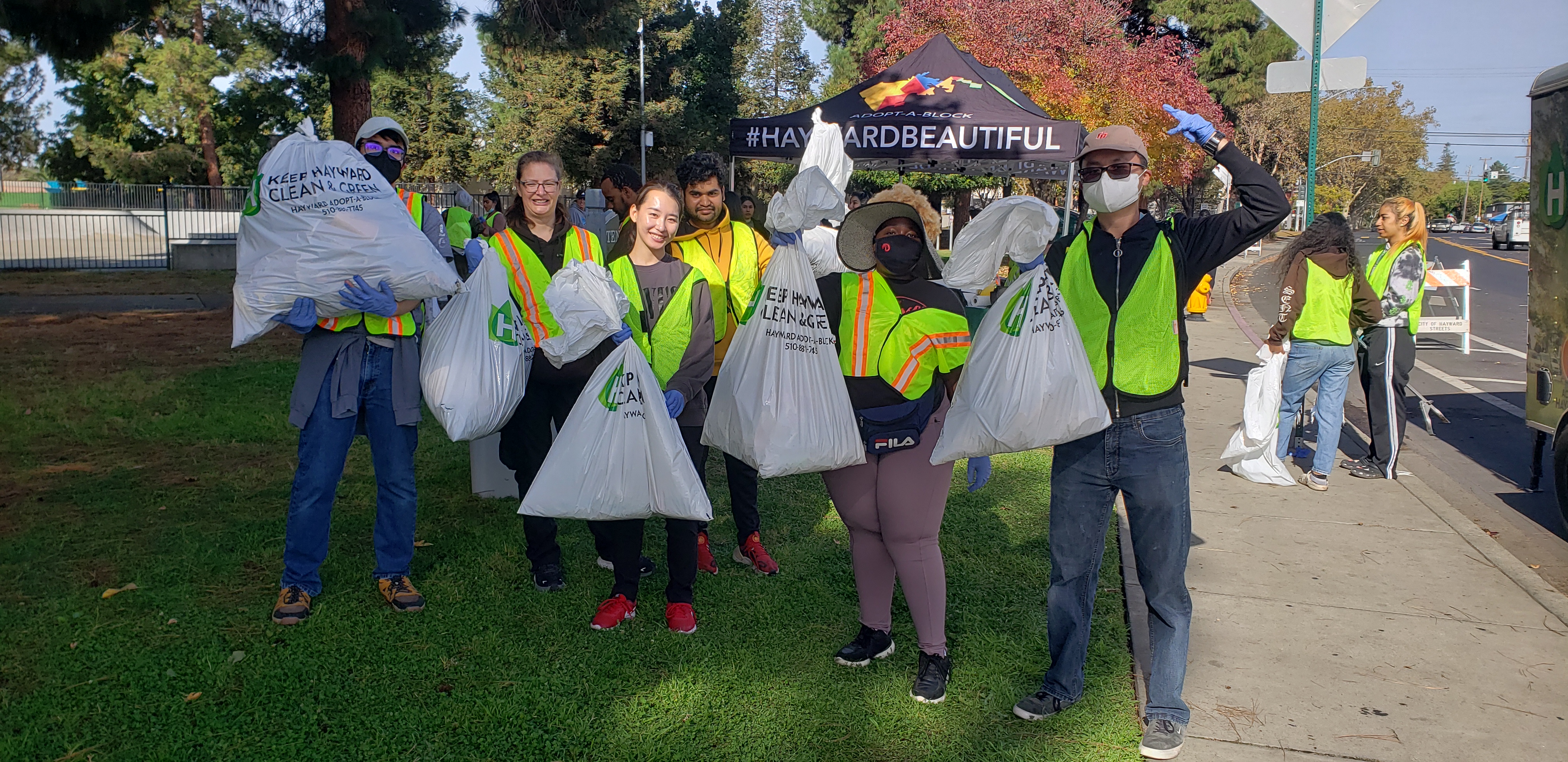 A picture of me and my classmates in yellow and red traffic vests carrying bags of trash in our hands as well as mechanical grabbers in our other hands.  We're at a shaded park in Hayward, California.