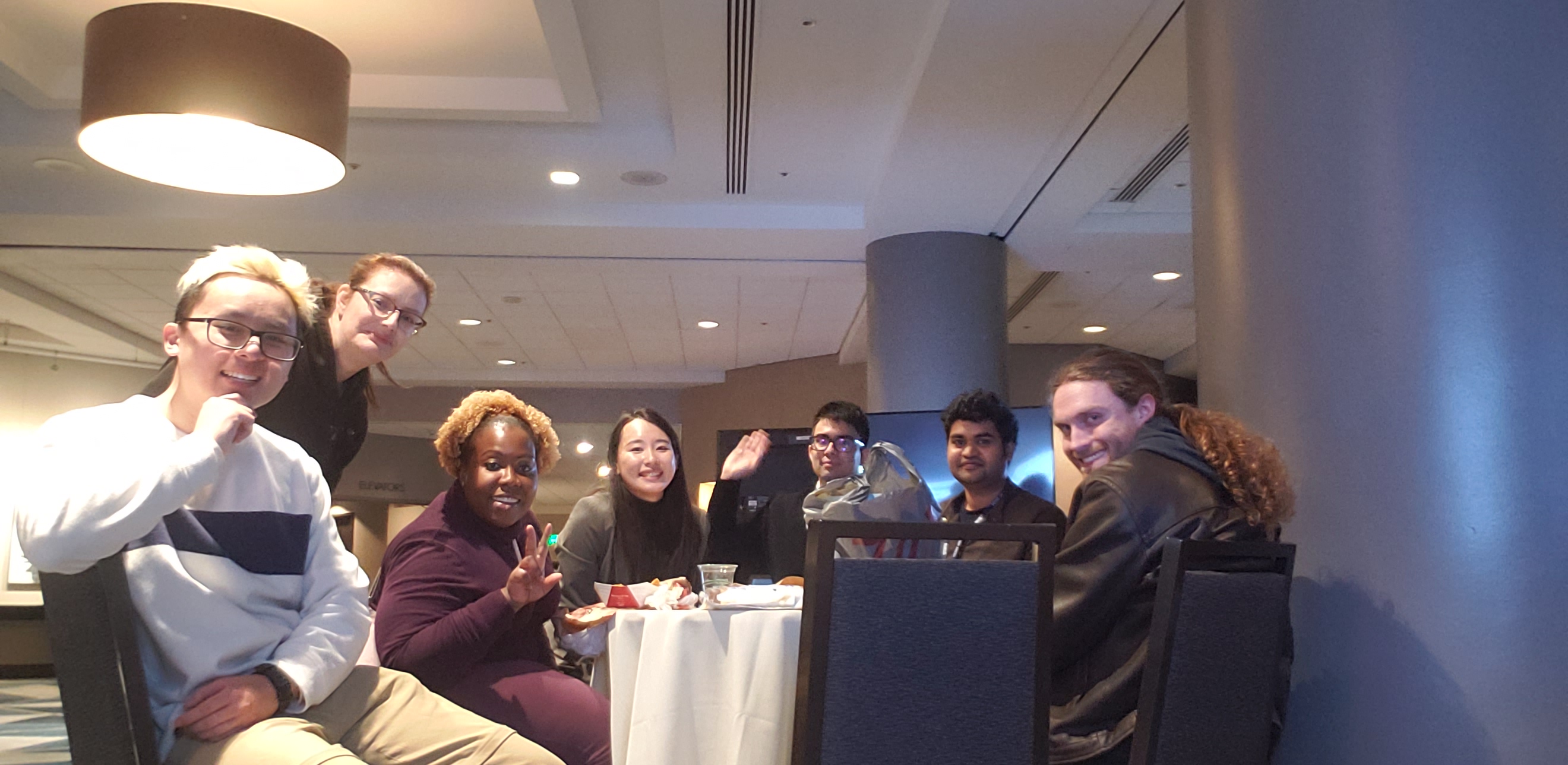 A picture of me and my classmates/friends posing at a table in the middle of an empty gathering hall in a hotel at night.