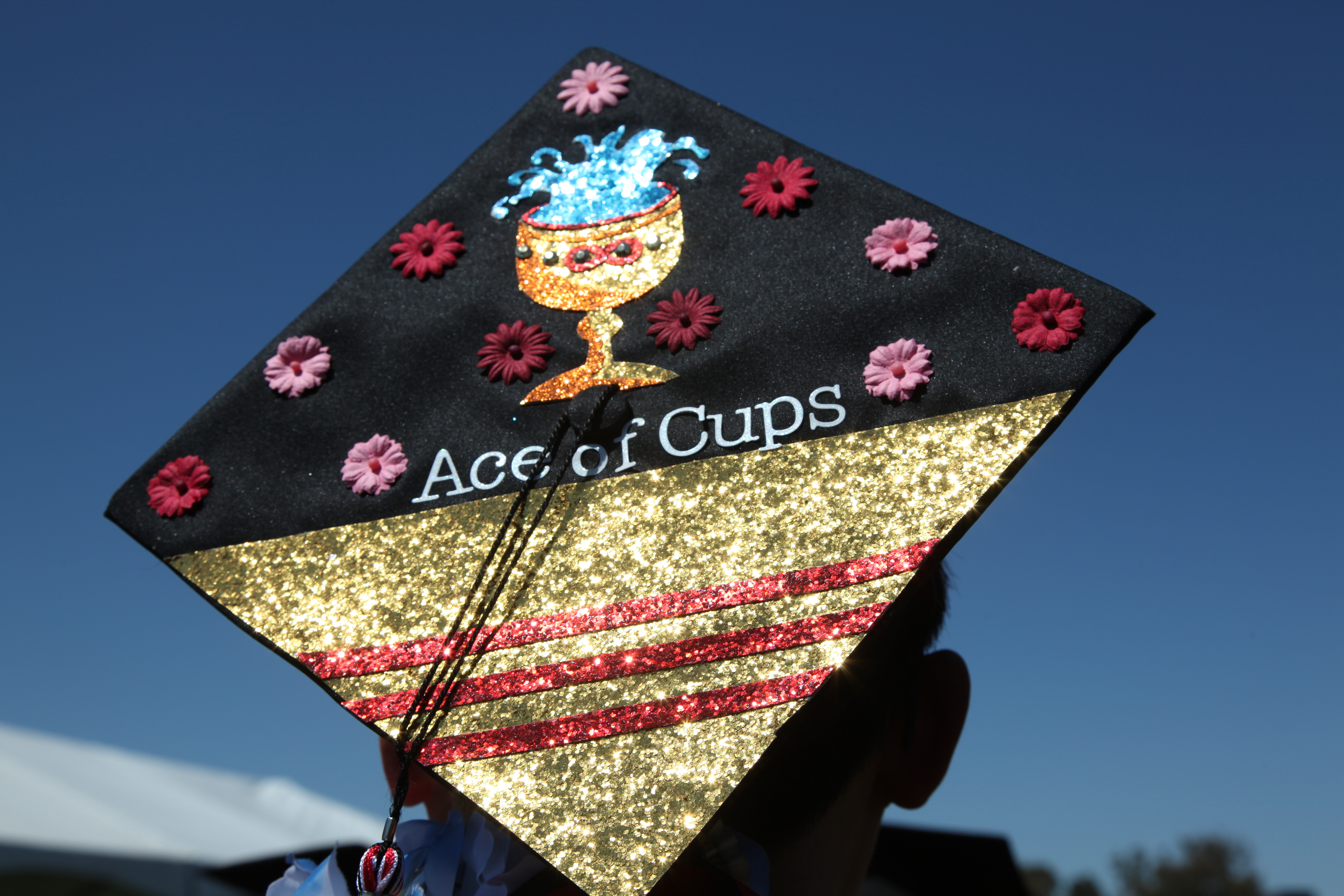 The back of my head featuring my black graduation cap, which contains a picture of a golden cup with water falling out and a red infinity symbol in the middle of the cup.  The top half of the black graduation cap is decorated with red and pink flower tops sprinkled around.  The middle of the cap below the golden cup reads, 'Ace of Cups'.  The bottom half of the black graduation cap is the flag of South Vietnam, featuring a golden background with three red stripes in the middle.