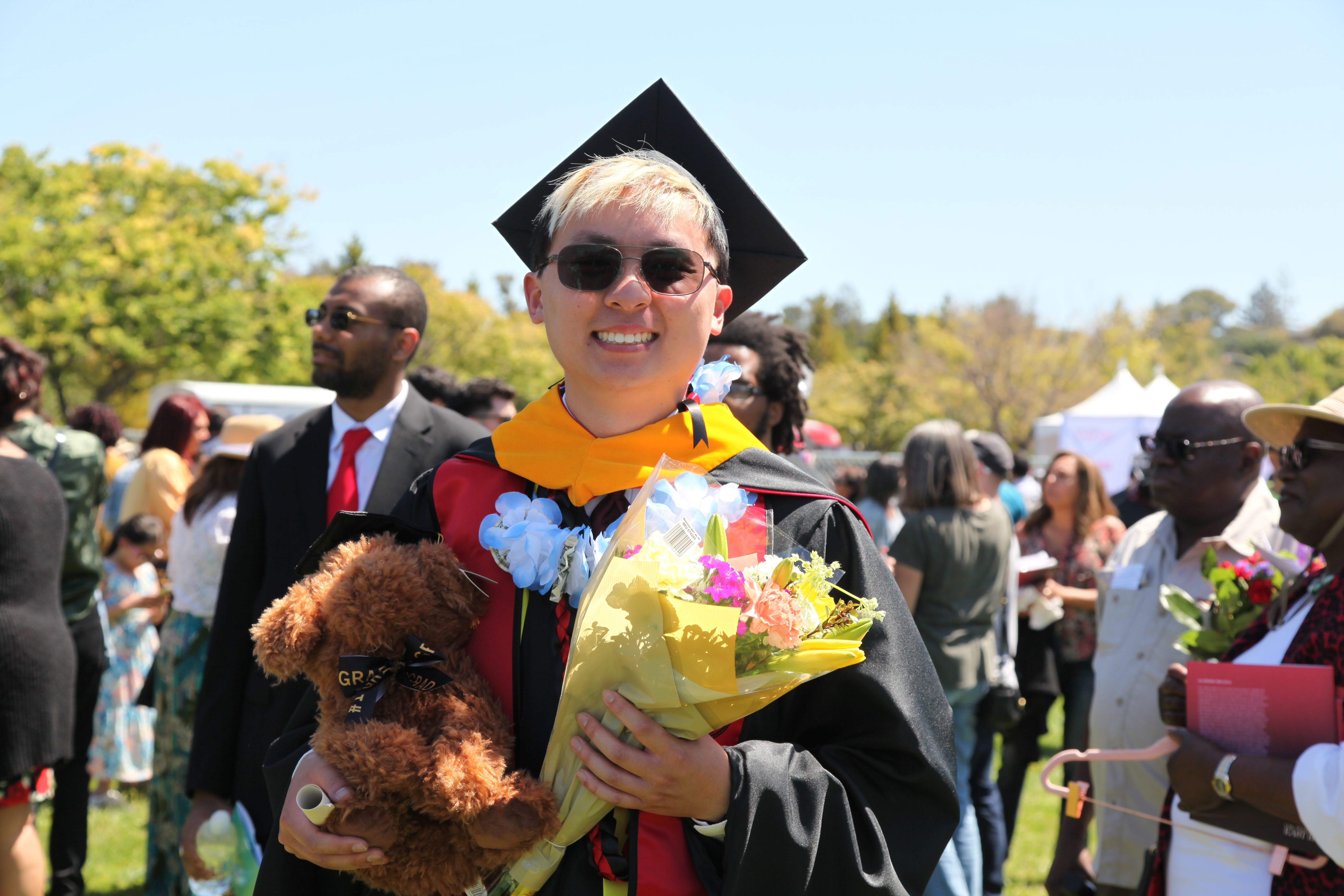 Ken Vu wearing a graduation gown while carrying a bouquet of flowers, a scroll, and a stuffed animal dog in his arms.
