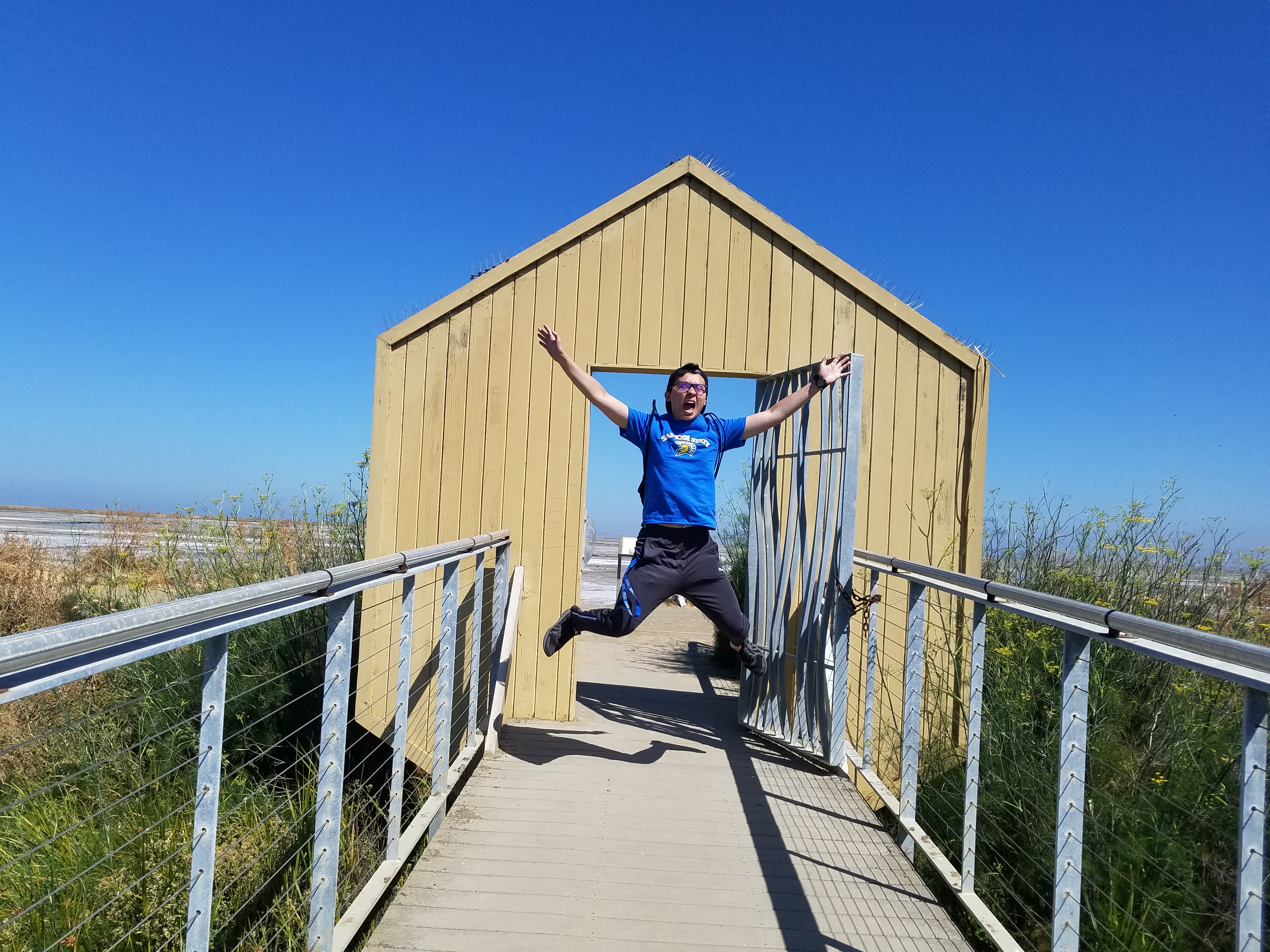 A picture of me jumping on a wooden walkway with guardrails in front of a door frame on top of the walkway across a grassy creek.  There is a clear blue sky overhead and bright sunlight across these vast saltlands.