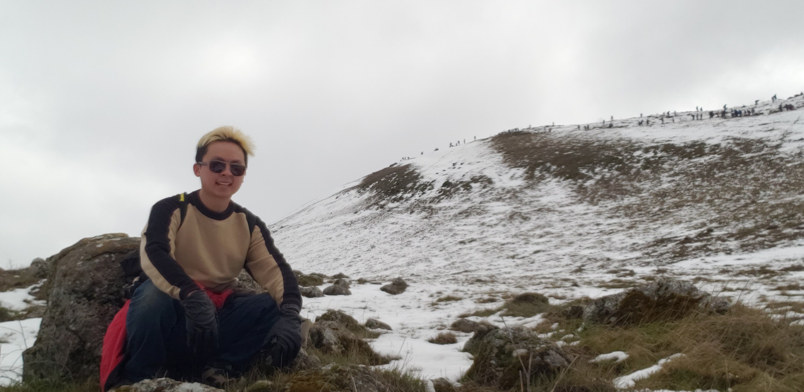A picture of me with sunglasses, a brown sweater, and black gloves crouched down in front of a rock.  I'm near the top of a snowy hill under cloudy conditions in the afternoon.