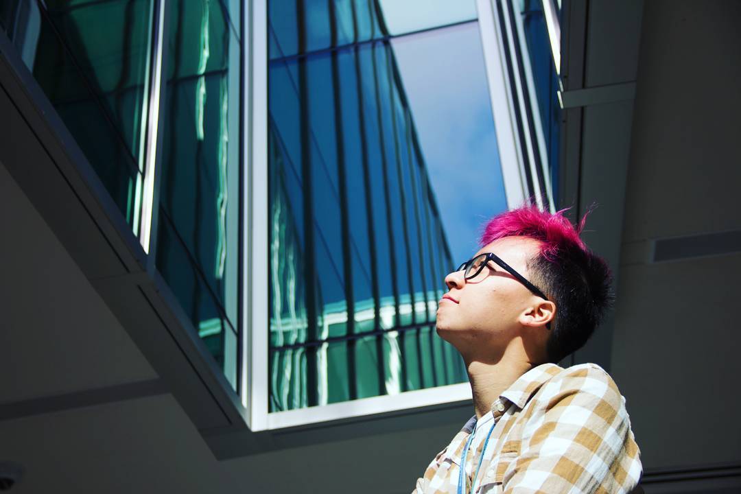 A picture of me with the top of my hair dyed a red-pink color with a white and light brown patterned flannel as I look up outside at a clear blue sky while I stand near an opening at building with glass windows.