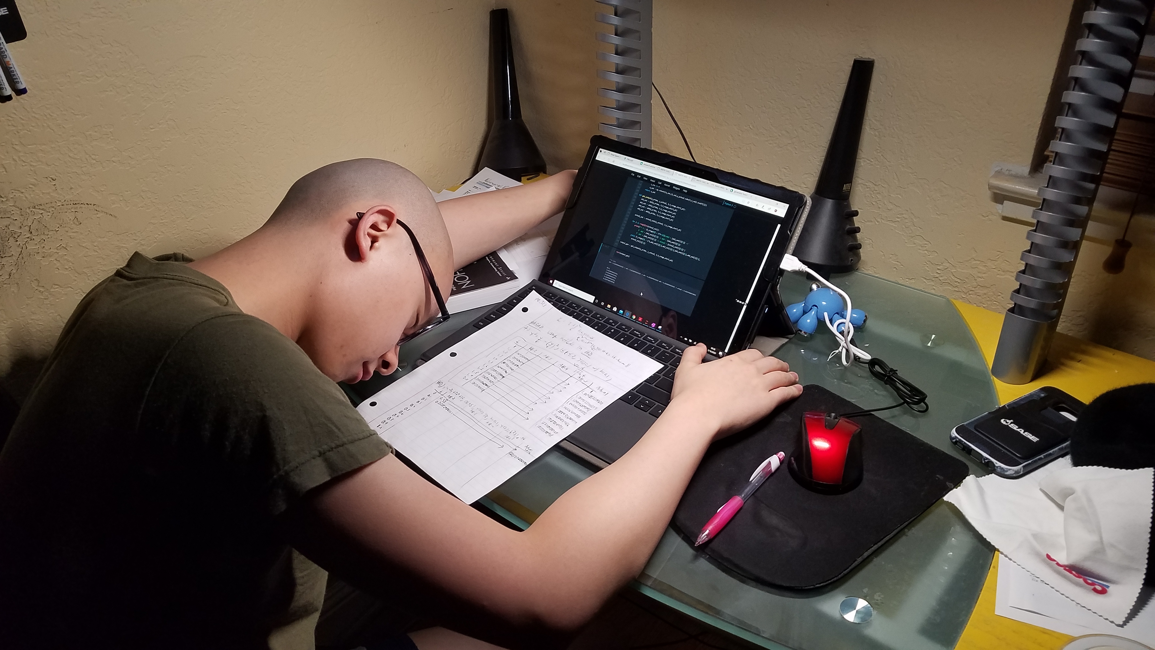 Ken Vu with his face down on his dimly lit desk at night while he has his laptop on and a class homework sheet in front of him.