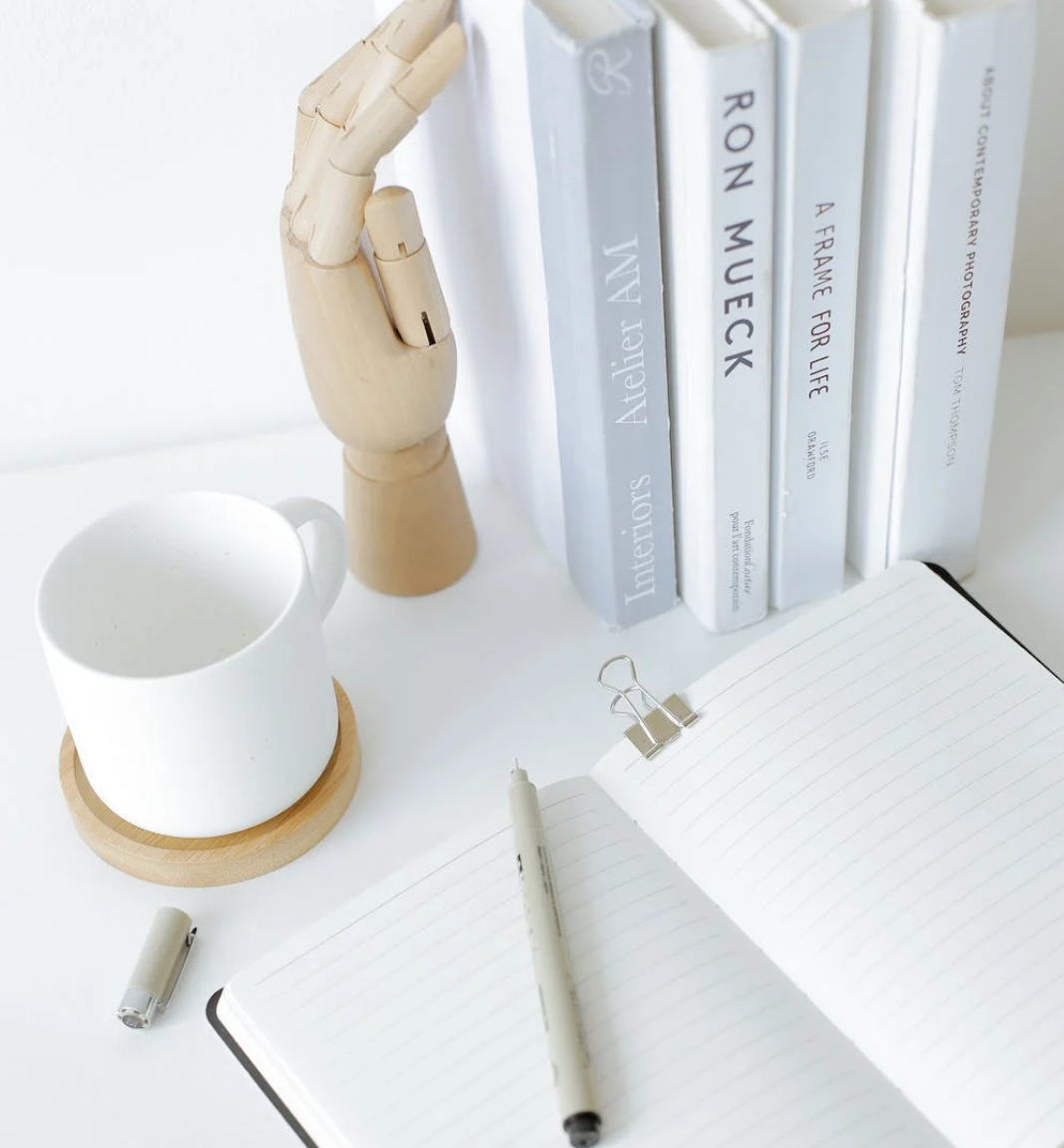 A picture of an open notebook with a pen on top with an upright stack of books in front of it - all of which are on a desk.
