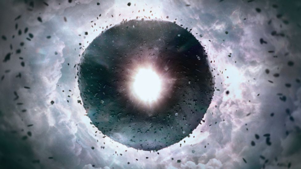 A picture of a dark grey background of clouds with a black bagel-shaped object with sprinkles on it and a bright hole in the middle with white light coming out  as seen in the movie 'Everything Everywhere All at Once'.