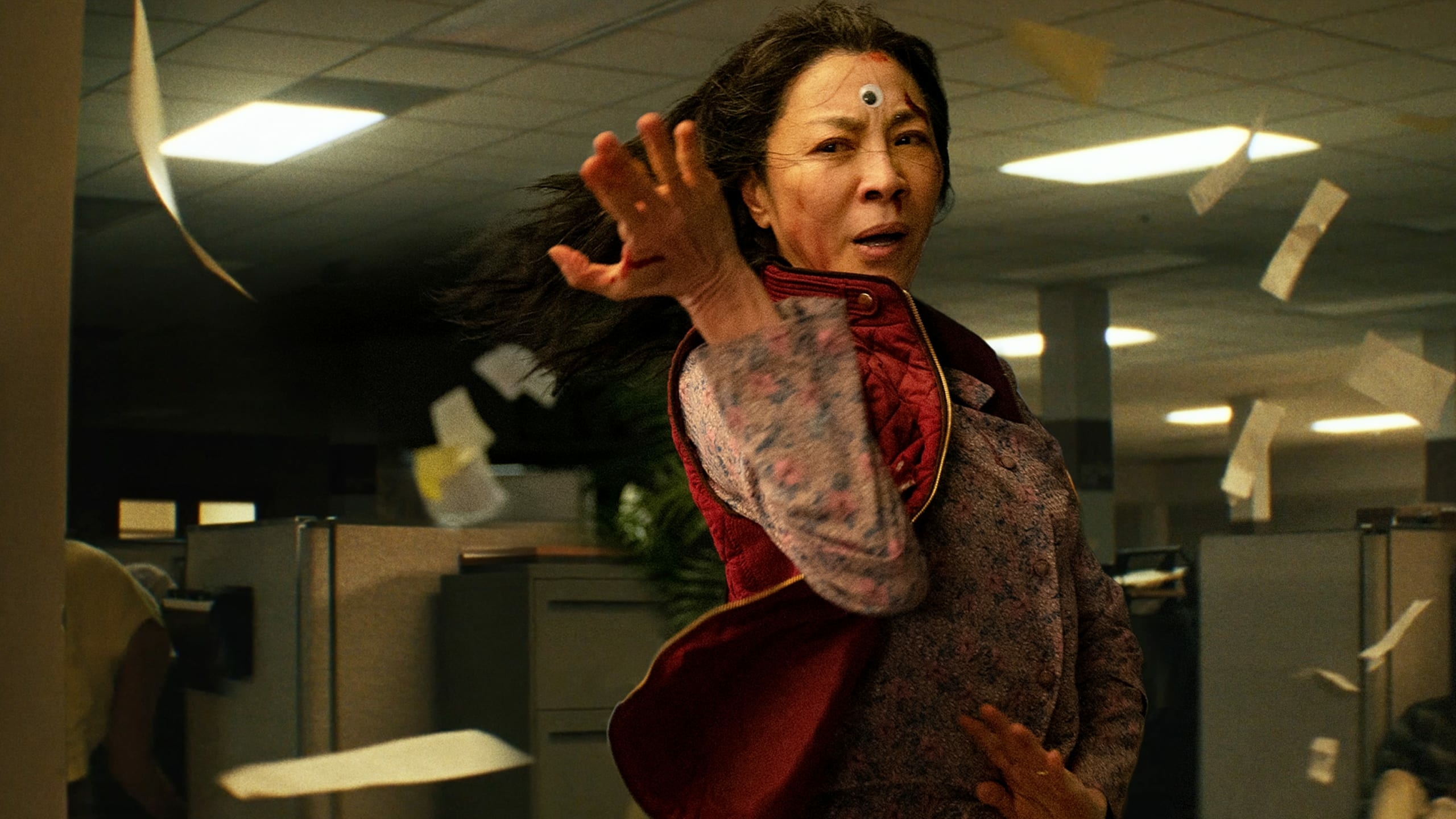 A picture of an Asian woman holding her hand out in an office cubicle setting in the background while she wears a googly eye on her forehead and papers fly all around her as seen in the movie 'Everything Everywhere All at Once'