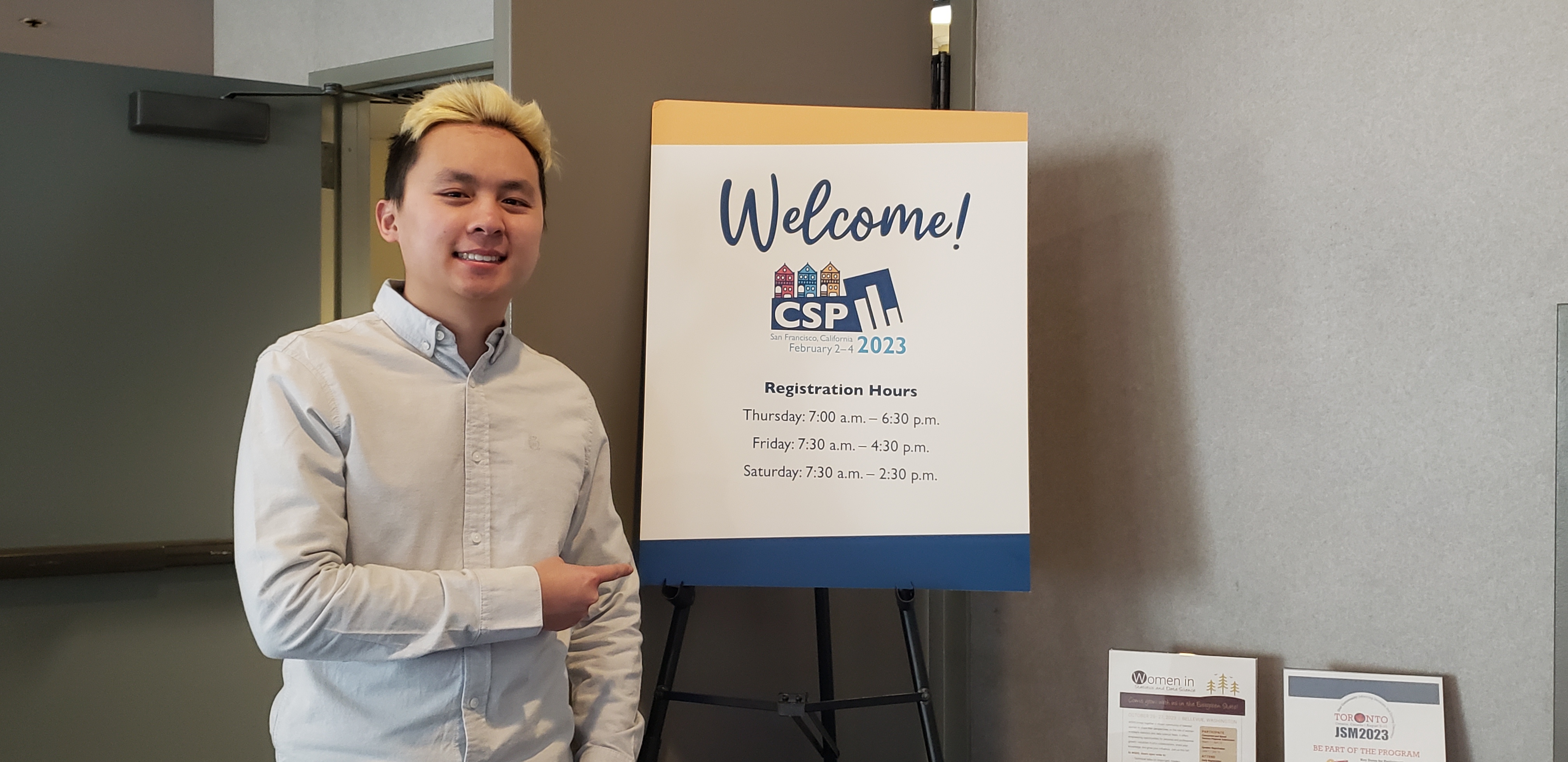 A picture of me in a white button up shirt standing next to a poster saying 'Welcome to CSP 2023' as I point to a poster.