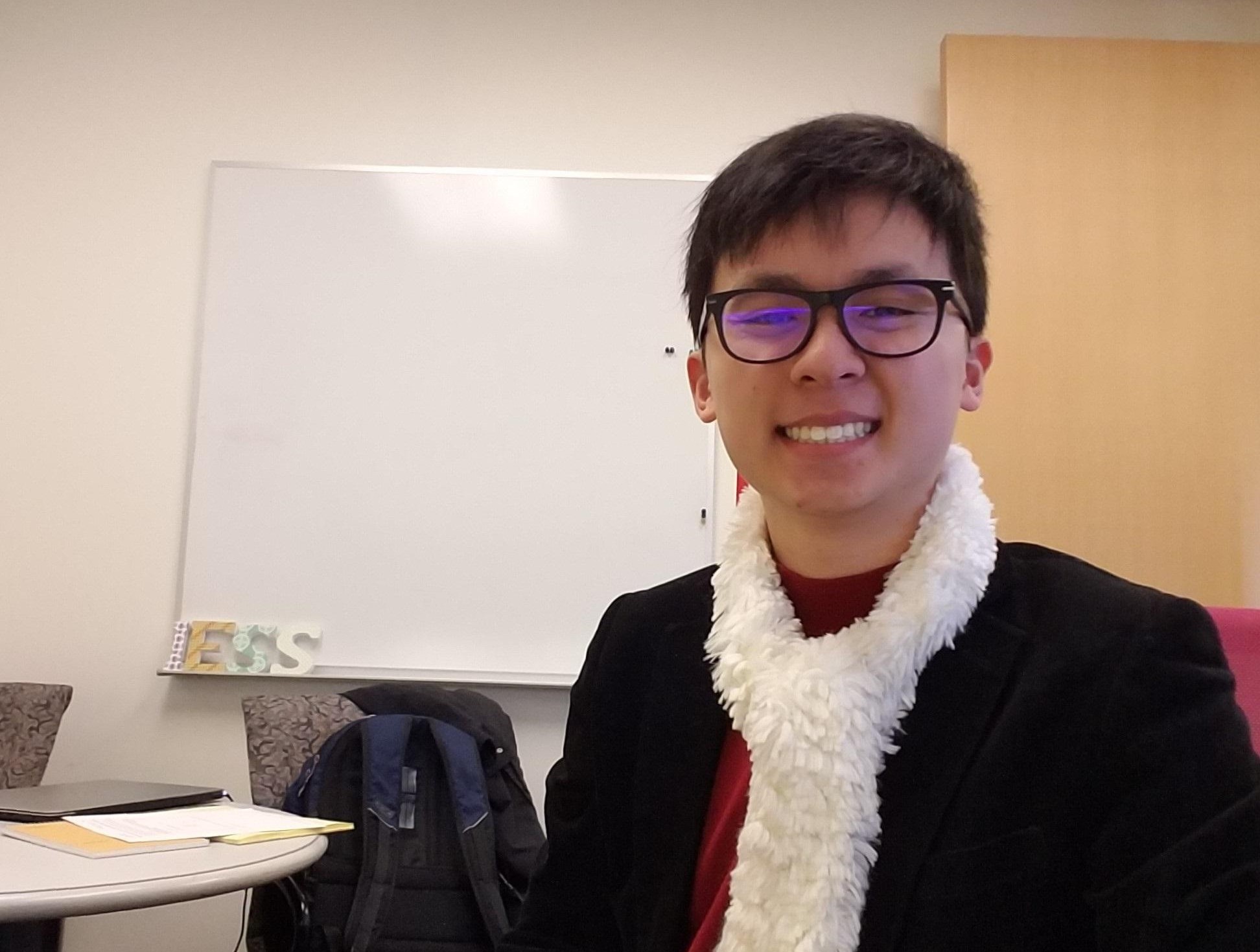 A picture of Ken Vu sitting in an office area with his white scarf around his neck, his red turtleneck, and his black coat on.