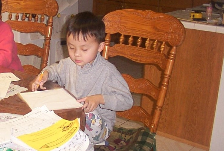 A photo of a young Ken as a child preparing to write in his notebook while seated at a dining room table.