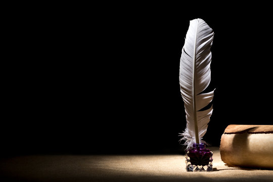 An illuminated spot in a dark room with quill standing upright while dipped in an inkwell next to a scroll.